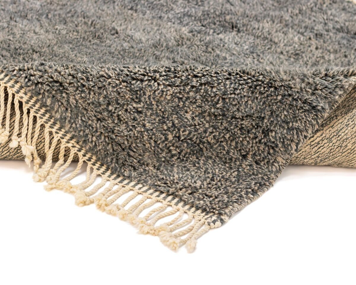 Close-up of a gray and beige hand-knotted rug with tassels on the edges, showing a corner fold that reveals the rugs woven underside. Its authentic Moroccan Rus design adds an artisanal touch to any space.