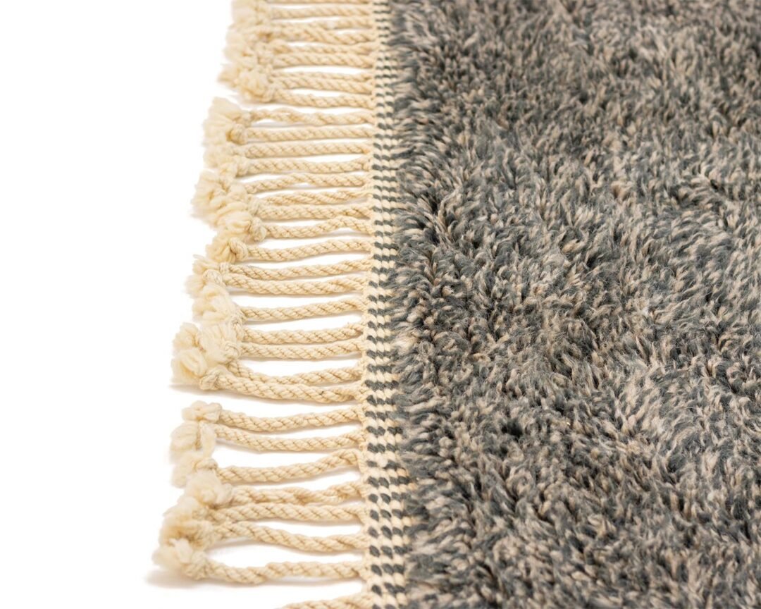 This close-up showcases an authentic Moroccan rug, featuring a textured gray surface with cream-colored fringes along the edge. The artisanal piece is hand-knotted, adding a touch of elegance and craftsmanship to any space.