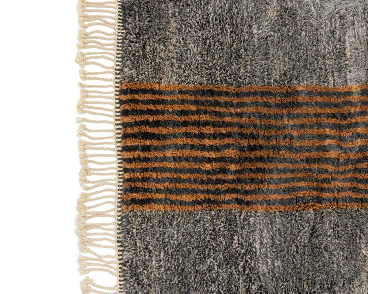 This artisanal rug showcases a fringe on the left, with a gray background and a central horizontal stripe pattern in vibrant orange and black tones. Hand-knotted craftsmanship adds to its unique charm.