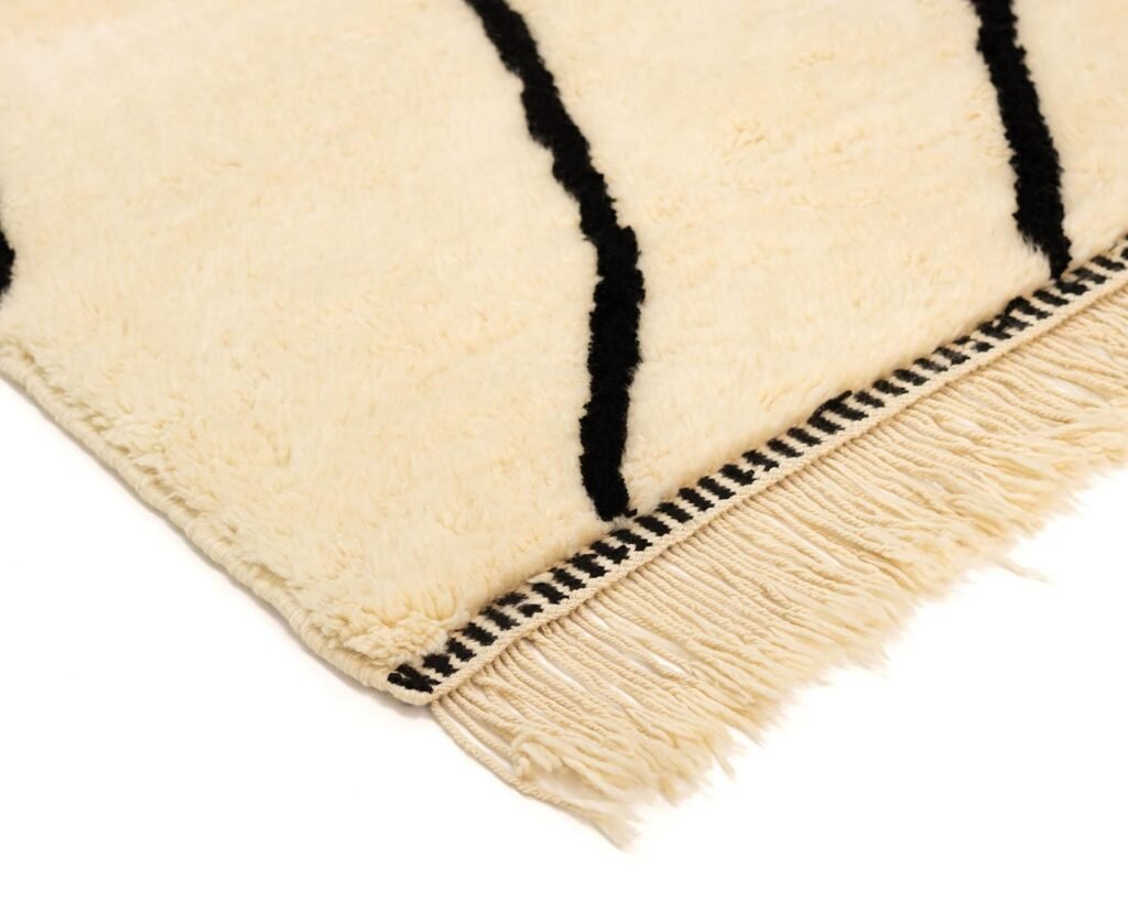 This cream-colored Moroccan rug features black linear patterns and fringed edges, showcasing artisanal charm in any space.
