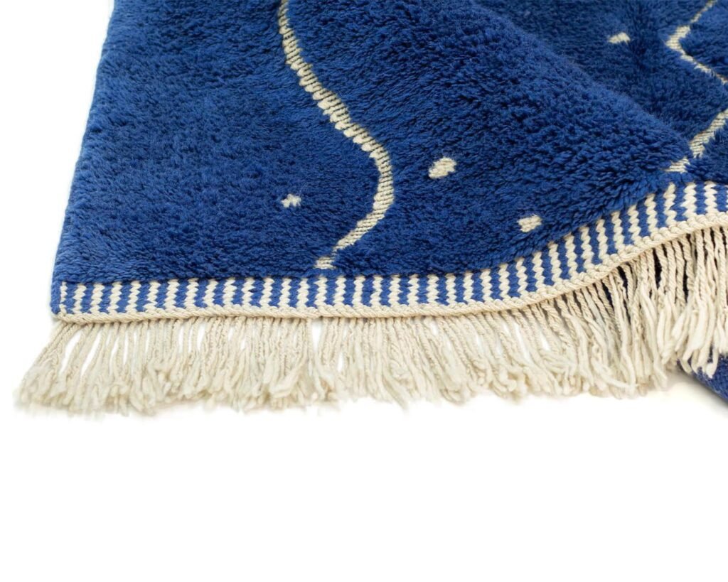A blue fringed towel adorned with white decorative patterns like dots and wavy lines is neatly displayed on a white surface.