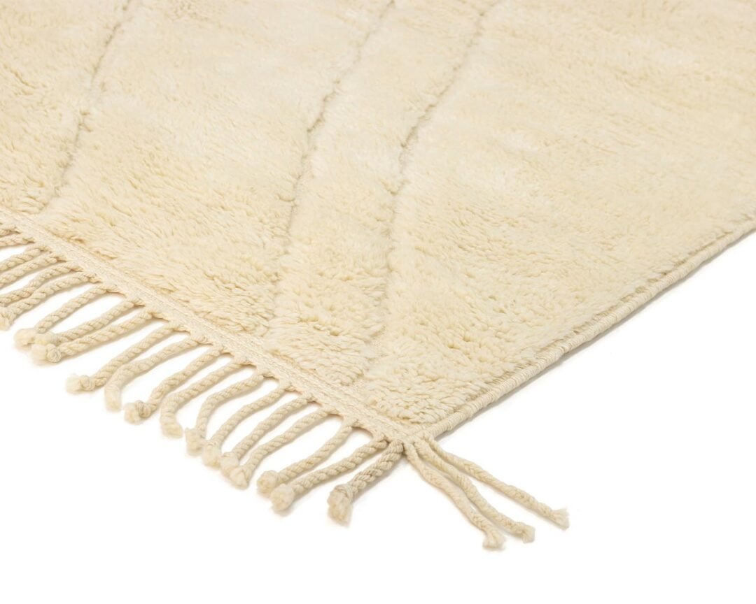 Close-up of an authentic Moroccan rug, hand-knotted with cream-colored fibers and fringe tassels on the edge, showcasing a textured, soft surface.