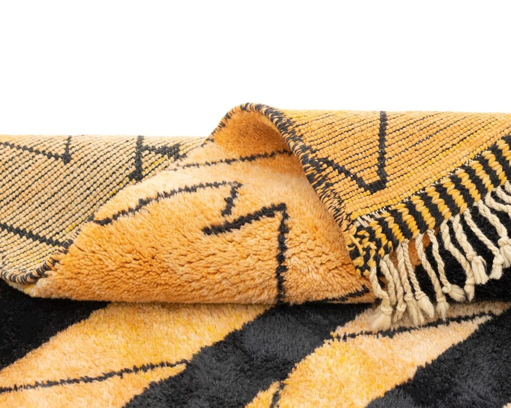 A folded, textured rug with a geometric pattern in yellow, orange, and black, featuring fringe on one edge.