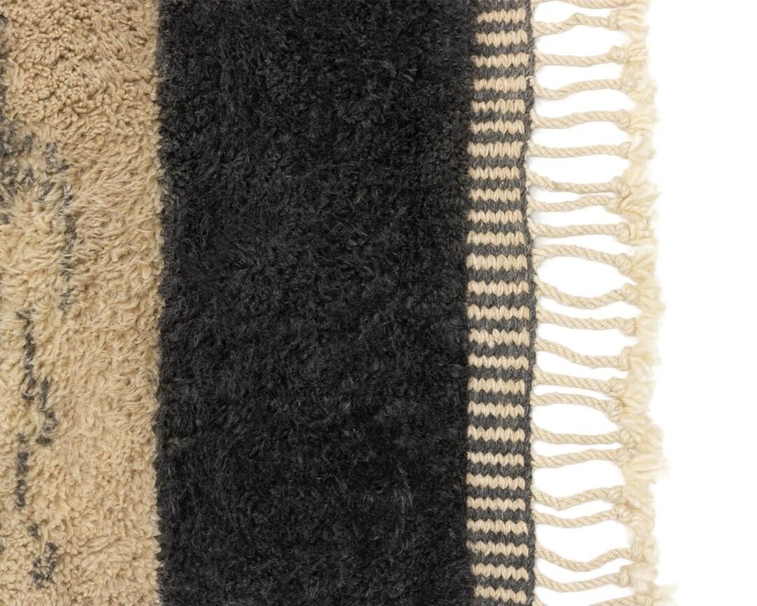 This image shows a close-up of a rug with vertical stripes in beige and black, adorned with fringed tassels on the right edge. Its intricate pattern adds a sophisticated touch to any space.