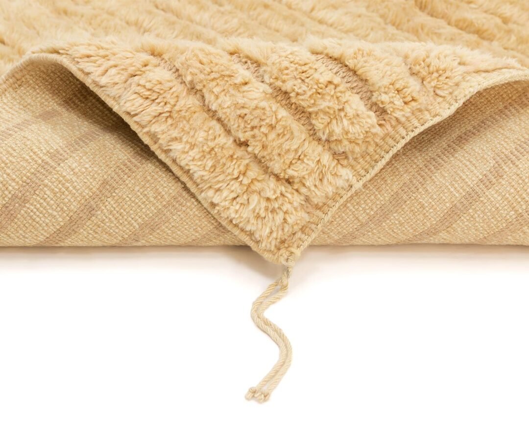 Beige luxury rug with a folded corner revealing its fluffy underside and a loose string. Crafted as part of our handmade home decor collection, it adds a touch of elegance to any space.