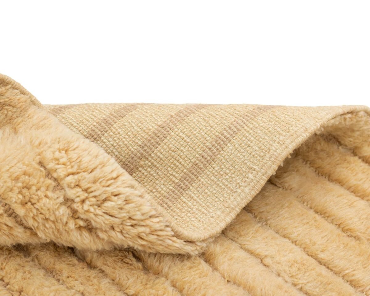 A luxurious, beige rug with a rolled corner reveals a striped underside. This hand-knotted masterpiece boasts a soft, fluffy appearance resembling fur, perfect for elegant home decor.