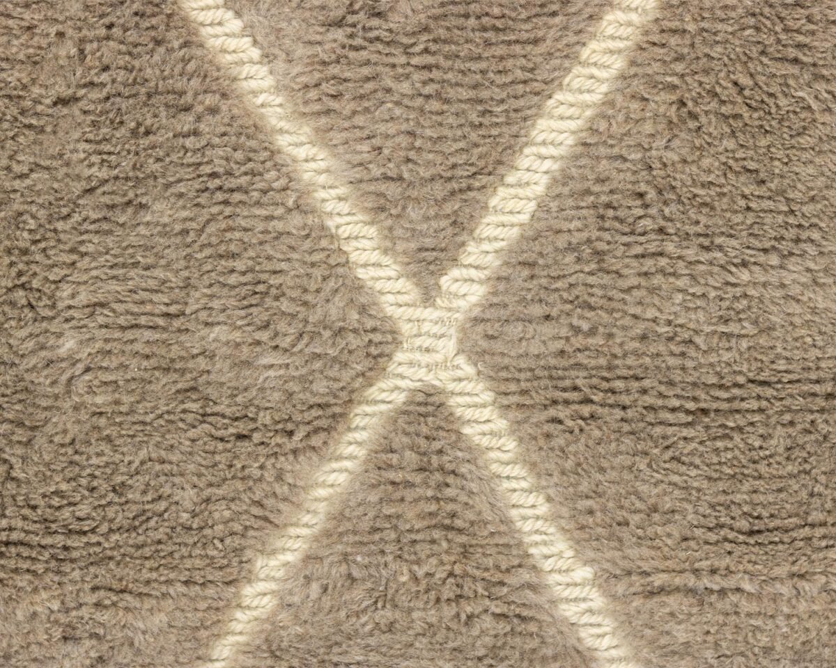 A close-up of a hand-knotted rug reveals a textured gray surface with a cream-colored crisscross pattern, showcasing the craftsmanship typical of authentic Moroccan rugs.