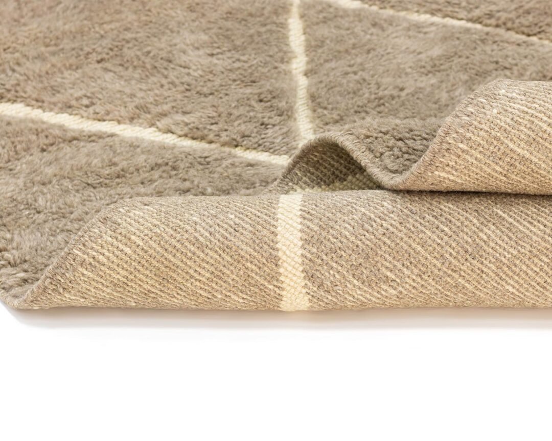 A close-up of an artisanal beige, textured rug with a white geometric line pattern, its edge slightly folded over on a white surface.