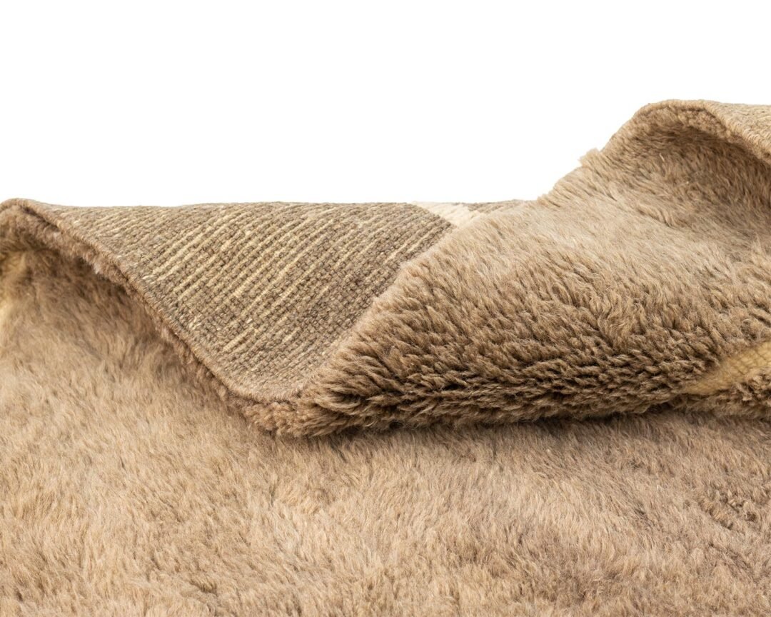 A close-up of a folded, light brown, fluffy faux fur with a woven underside highlights its texture and thickness.