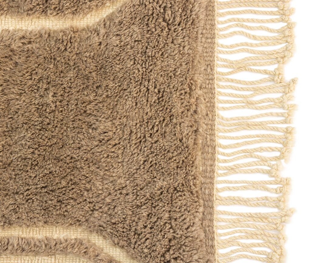 Close-up of a beige textured rug corner with fringed edges on a clear white background, adding an elegant touch to any room decor.