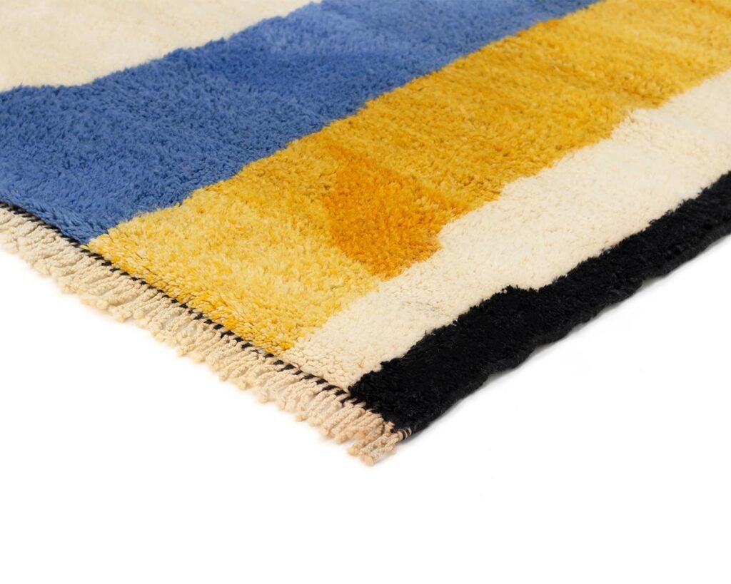 Close-up of an artisanal rug with blue, yellow, and black rectangular patterns on a white background. Hand-knotted with care, its vibrant colors and slight fringe speak to the skill of authentic Moroccan rug craftsmanship.