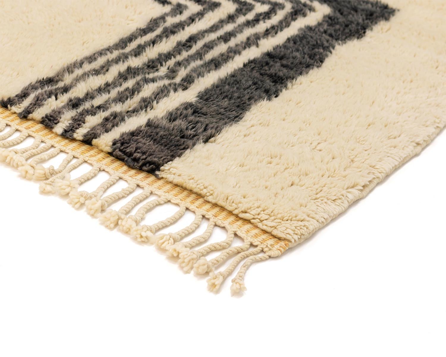 Close-up of a luxury beige and black woven rug corner with fringed tassels. The design features parallel lines forming an abstract pattern, reminiscent of a Moroccan rug style.