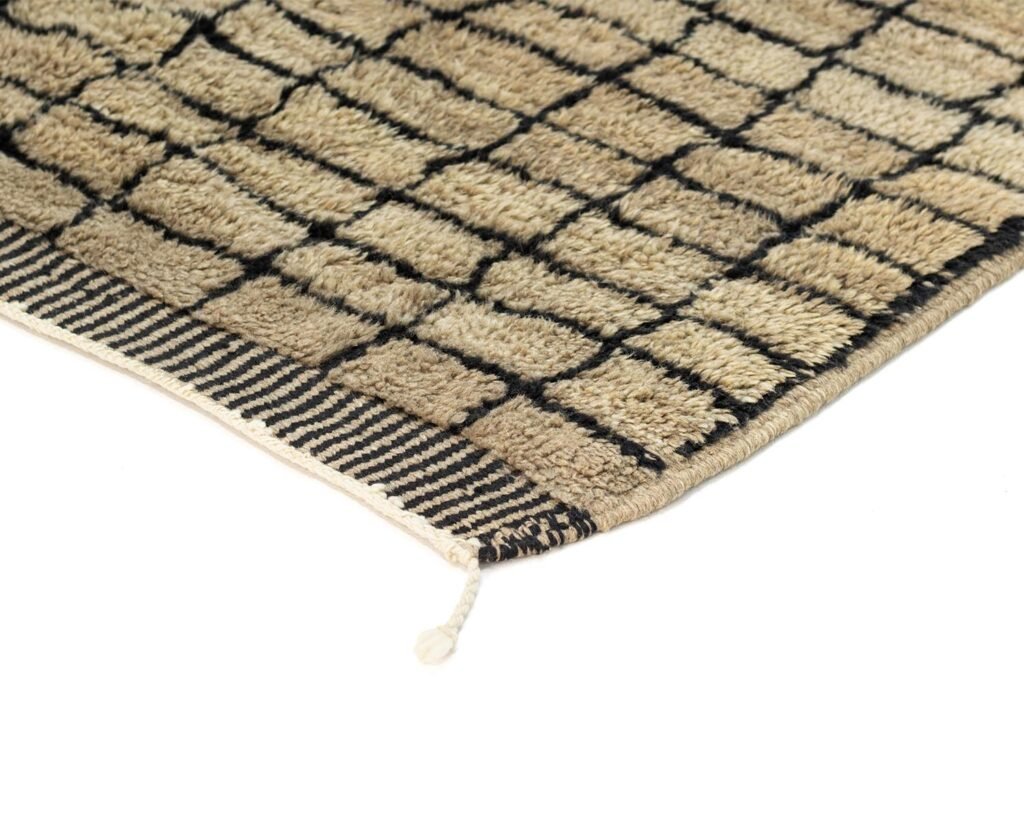 Experience the intricate details of this authentic Moroccan rug, hand-knotted with precision. Its beige surface features a grid pattern, bordered by a striking black striped edge. A unique touch is the loose thread extending from one corner, adding character to its elegant design.