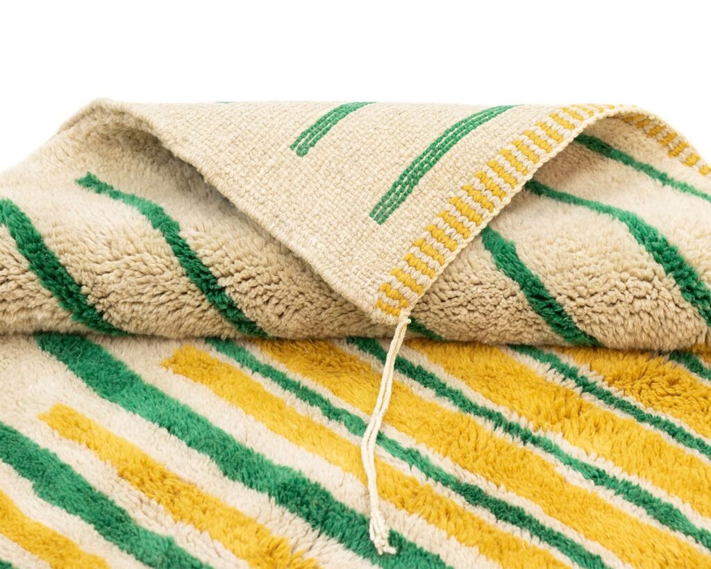 A folded rug with green and yellow stripes on one side and a beige woven pattern with green and yellow lines on the other side. A string is visible near the fold.