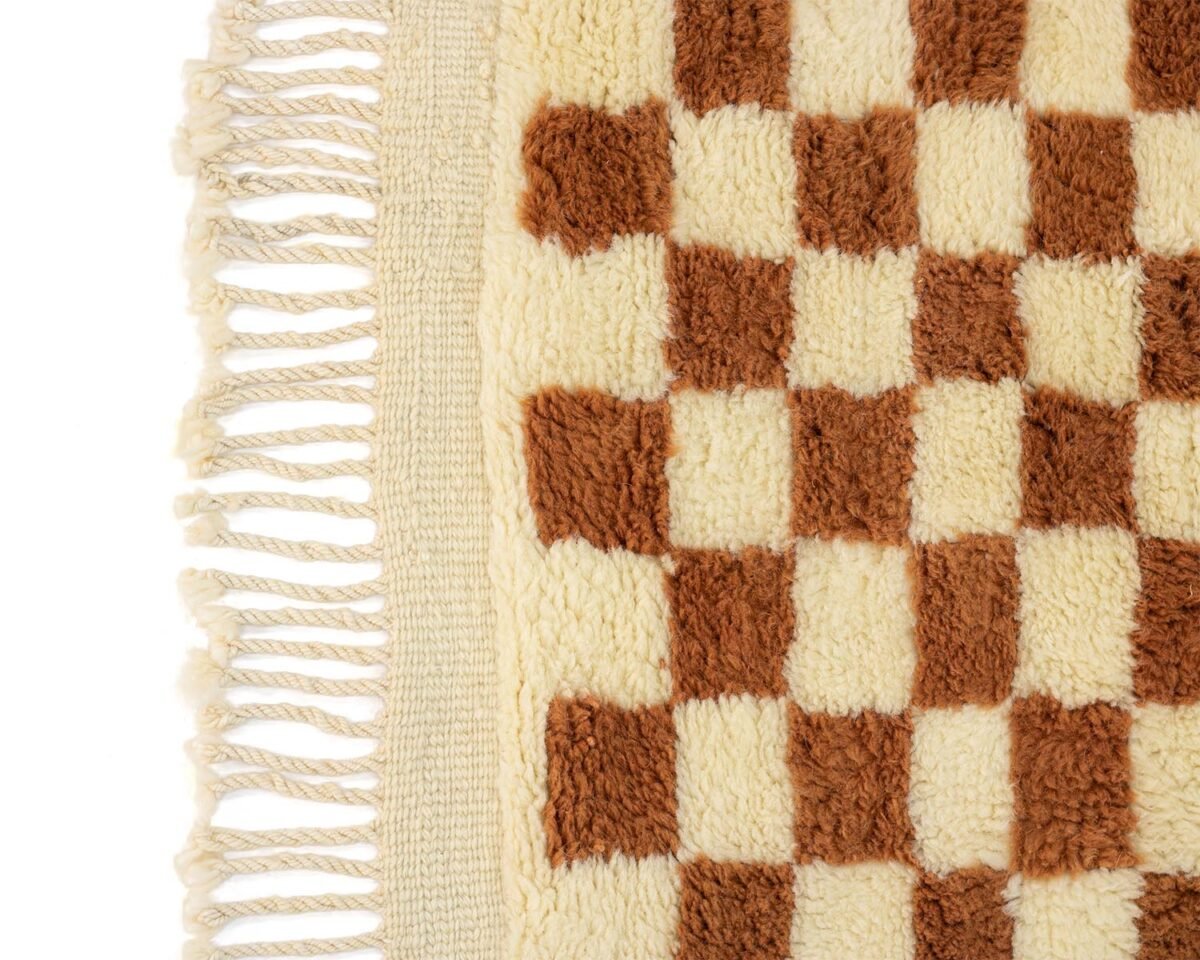 A detailed close-up of woven fabric showcases a brown and cream checkerboard pattern, complete with fringe along one side, capturing the artisanal craftsmanship and texture.