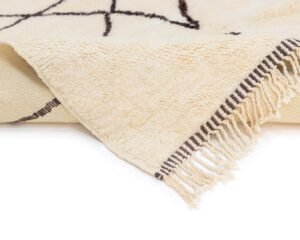A beige rug with intricate dark geometric patterns is artistically folded to reveal its fringed edges and the beautifully woven underside.
