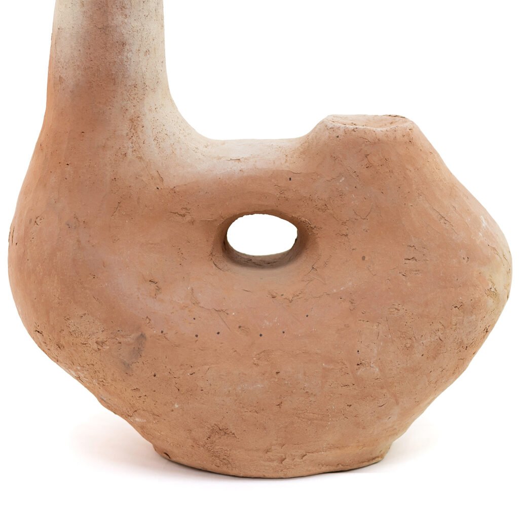 An unglazed Tamegroute pottery piece, this clay sculpture boasts an organic shape with a central hole. Its surface texture is rough and uneven, exuding a rustic charm with its light brown color.