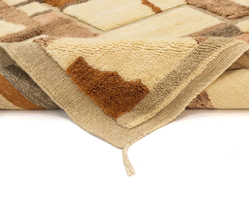 Close-up of a beige and brown geometric patterned hand knotted rug corner, slightly folded, with a loose thread on a white background.