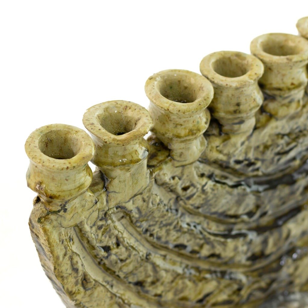 Close-up of a ceramic structure reminiscent of Tamegroute pottery, featuring a row of small, cylinder-shaped openings on a textured surface. The glaze is mottled with green and brown tones, suggesting an organic, earthy aesthetic.