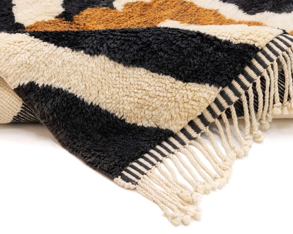 A close-up of a woven textile featuring black, beige, and brown abstract patterns with fringed ends resting on a flat surface. The intricate design and neutral tones add an artistic touch to any space.