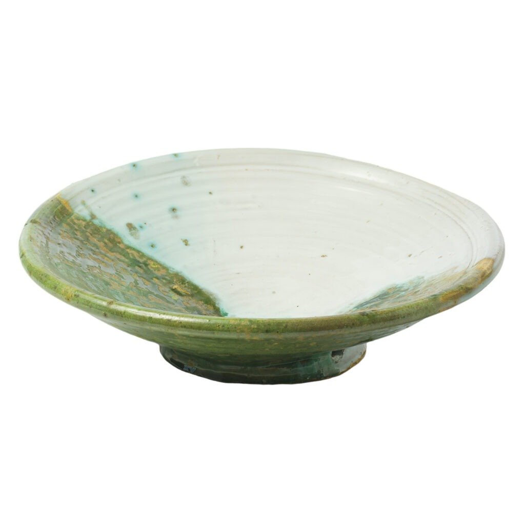 A shallow, round ceramic bowl with a wide rim, featuring a glossy green and white glaze characteristic of Tamegroute pottery, rests on a small pedestal base.