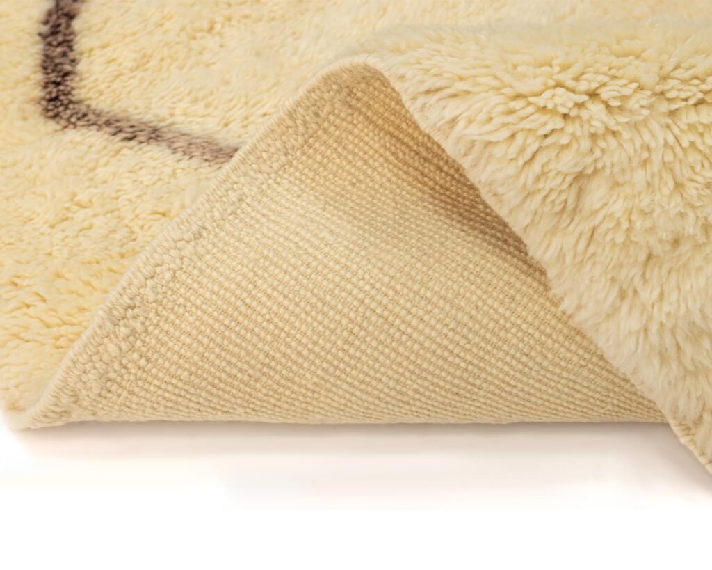 A corner of the beige, fluffy artisanal rug is folded over, revealing its textured underside, showcasing the craftsmanship of authentic Moroccan rugs.
