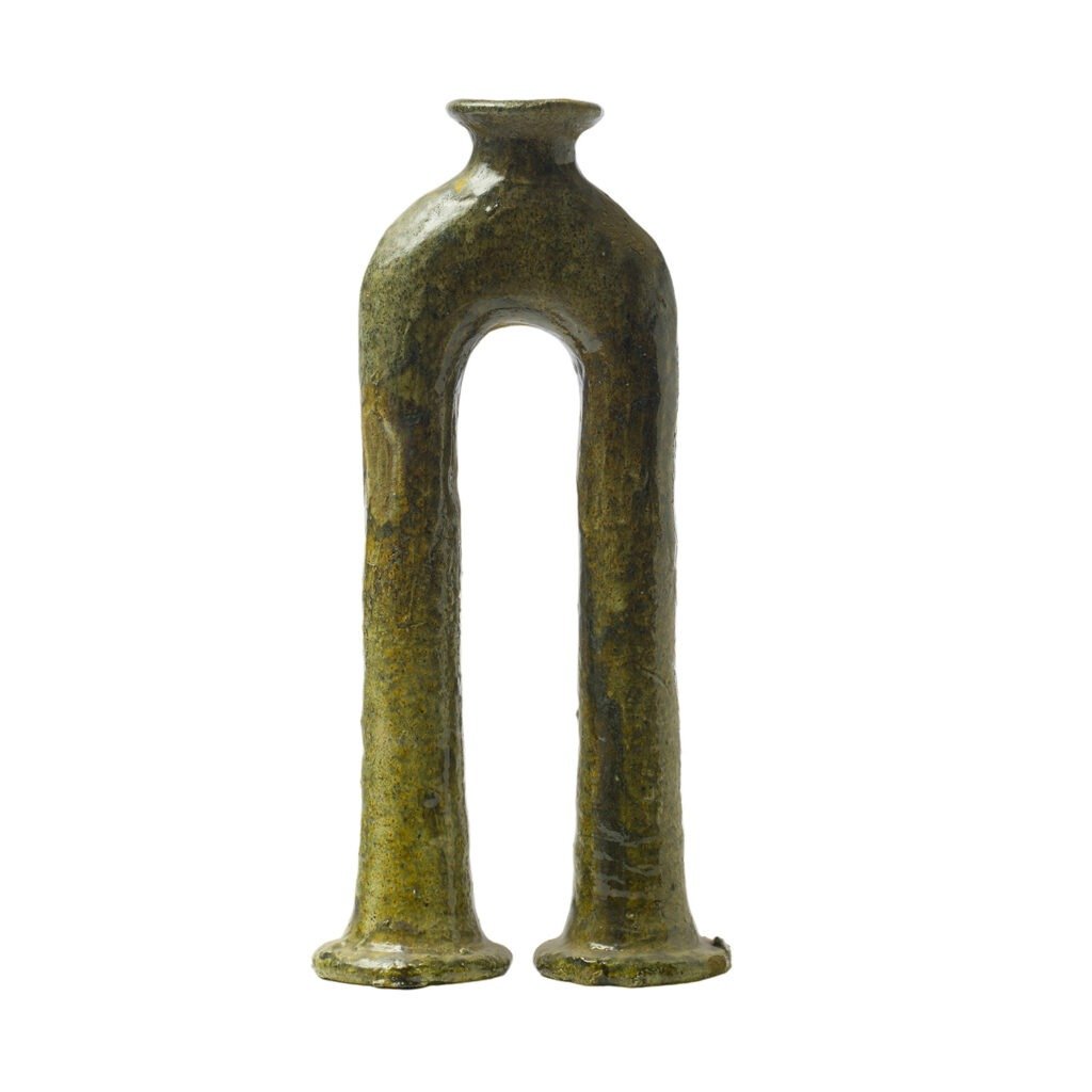 A tall, abstract ceramic vase with a unique arch shape and two legs features a textured, mossy green surface reminiscent of Tamegroute pottery.