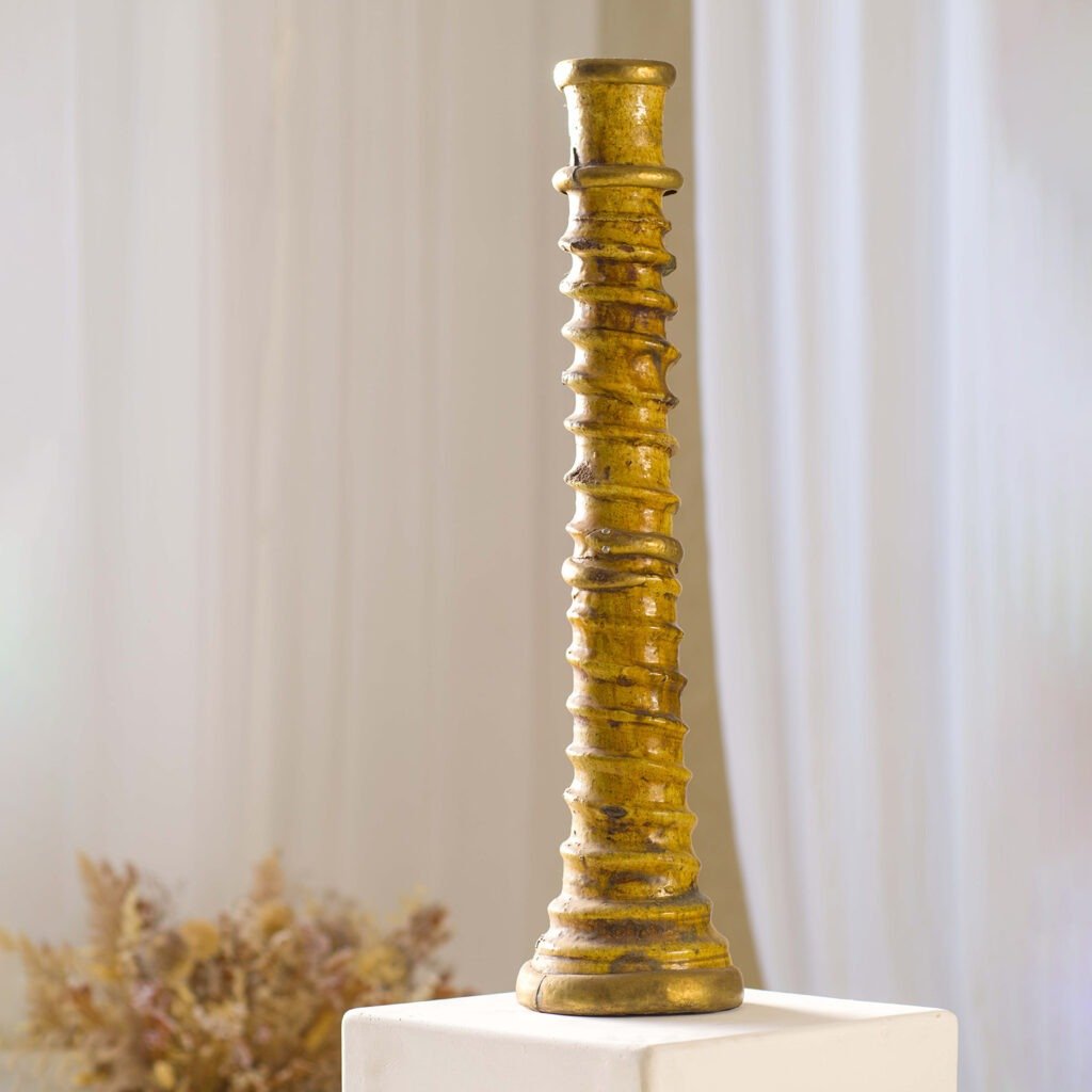 A tall, gold, spiral-patterned candlestick stands on a white pedestal against a blurred backdrop of dried flowers, embodying elegance and sophistication.