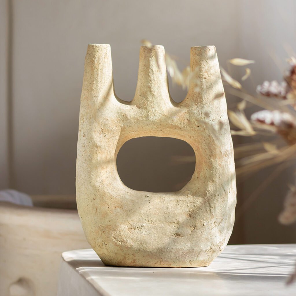 A textured, three-pronged beige ceramic vase with a large central hole, sitting on a white surface in soft, natural light. Elegant dried foliage provides a serene backdrop, adding to the artistry of this simple yet striking piece.