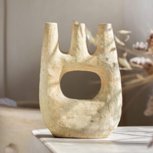 A textured, three-pronged beige ceramic vase with a large central hole, sitting on a white surface in soft, natural light. Elegant dried foliage provides a serene backdrop, adding to the artistry of this simple yet striking piece.