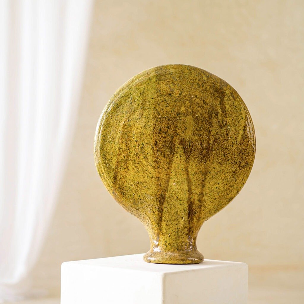 A round, flat-topped ceramic vase with a textured greenish-brown finish sits elegantly on a white pedestal against a softly blurred, neutral background.