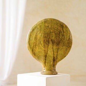 A round, flat-topped ceramic vase with a textured greenish-brown finish sits elegantly on a white pedestal against a softly blurred, neutral background.