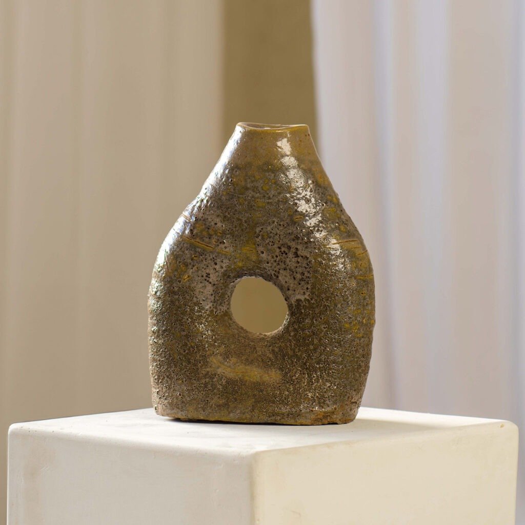 A brown, textured ceramic vase with a central circular hole sits elegantly on a minimalist white pedestal.