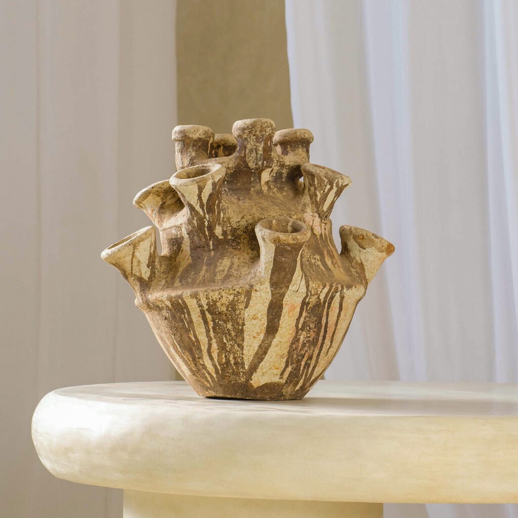 A textured, multi-spouted ceramic vase with a beige and brown pattern is elegantly placed on a light-colored table, accentuating the tranquil ambiance of white curtains in the background.