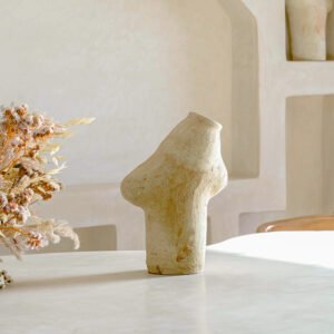 A serene, softly lit room showcases an abstract stone vase with dried flowers on a white table, complemented by sculpted wall shelves that add a touch of elegance.