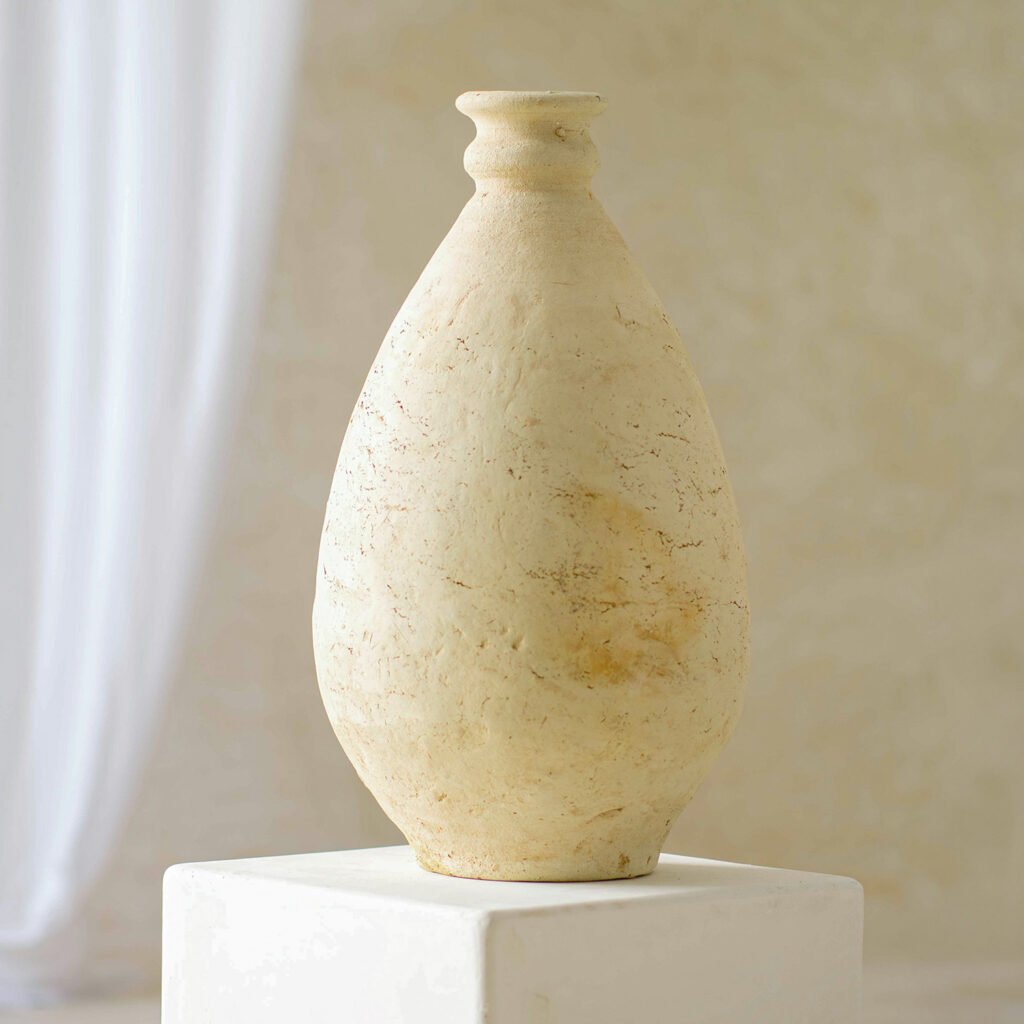 A tall beige ceramic vase with a narrow neck, textured surface, and a rounded body is elegantly displayed on a white pedestal against a neutral background, adding an artistic touch.