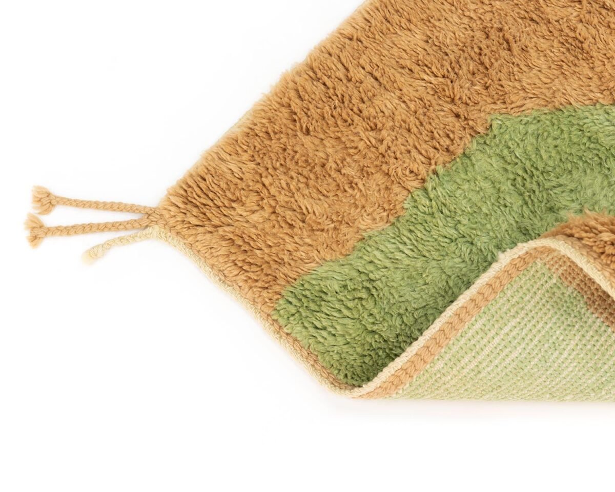 A close-up of a corner of a hand-knotted rug with tan and green sections showcases its luxurious texture and elegant tassels. The handmade home decor piece is partially folded, revealing its intricate underside.