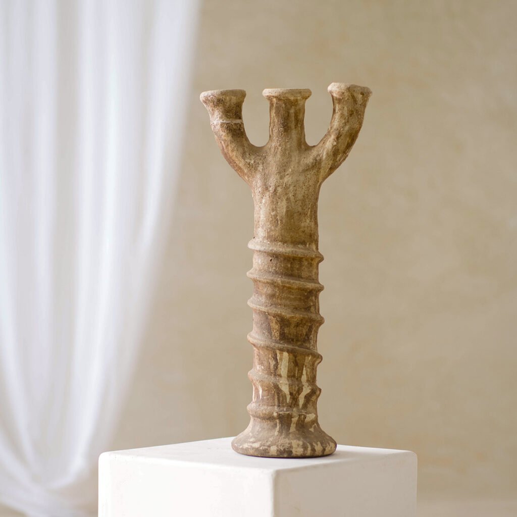 A clay sculpture with three upward-extending prongs and a ridged column stands on a white pedestal against a soft, neutral backdrop. Its intricate design captures the essence of artistic expression, highlighting the beauty of raw clay.