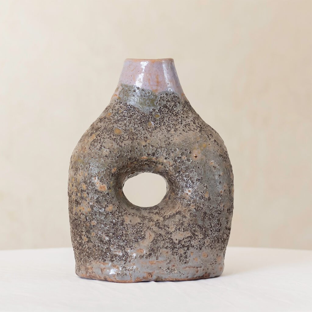 A textured, irregularly shaped glazed Tamegroute pottery vase with a circular hole in the center is displayed on a white surface against a neutral background.