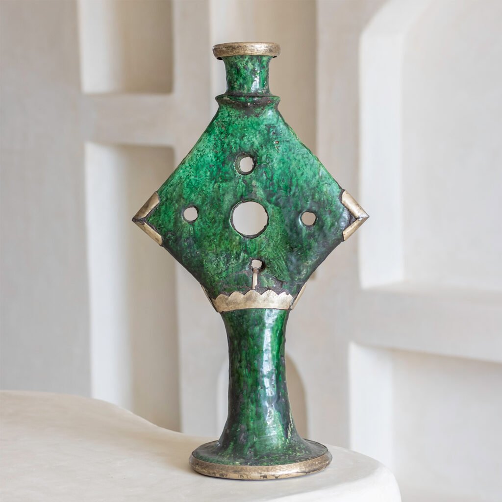 A green, intricately designed ceramic vase with a diamond shape and circular cutouts is displayed on a rounded base against a serene setting. Its artistic detail adds an elegant touch to the surrounding decor.