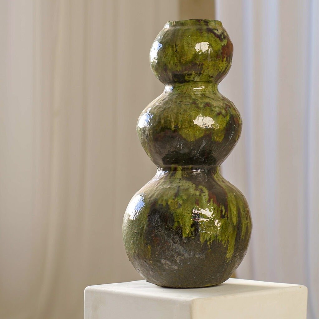 A glossy, emerald three-tiered ceramic vase rests on a white pedestal against a neutral curtain backdrop, exuding timeless elegance.