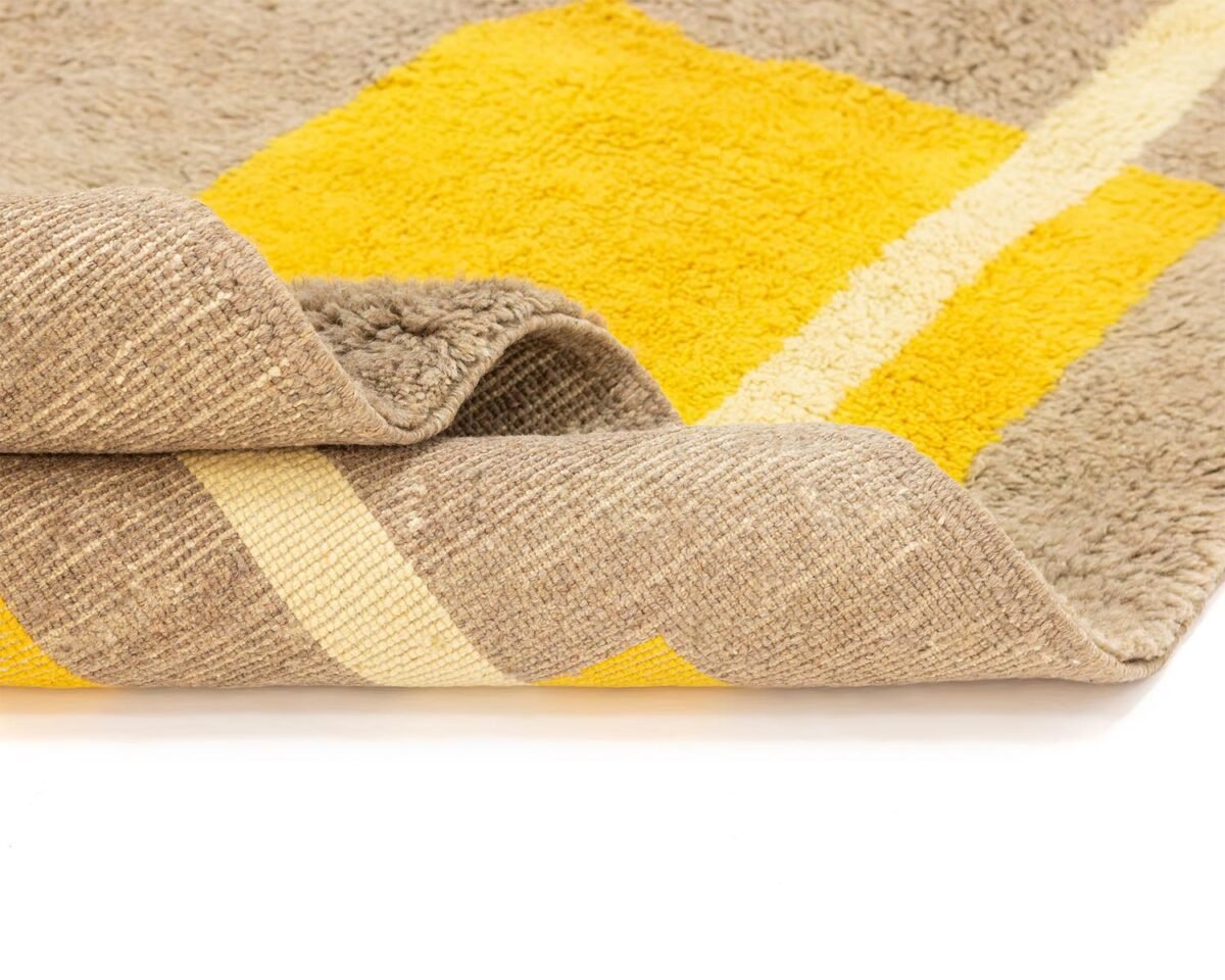 A corner of the hand-knotted rug is folded, revealing a yellow square pattern atop a textured surface and a beige underside adorned with yellow and white stripes. This luxury rug adds an elegant touch to any room.