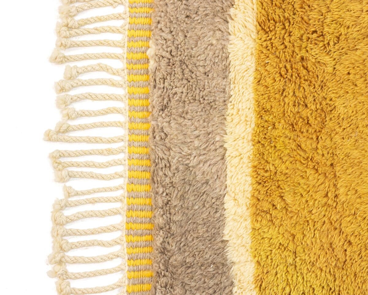 Close-up of a luxury multi-colored Moroccan rug with varying shades of yellow and brown stripes, featuring tasseled fringes on the left side.