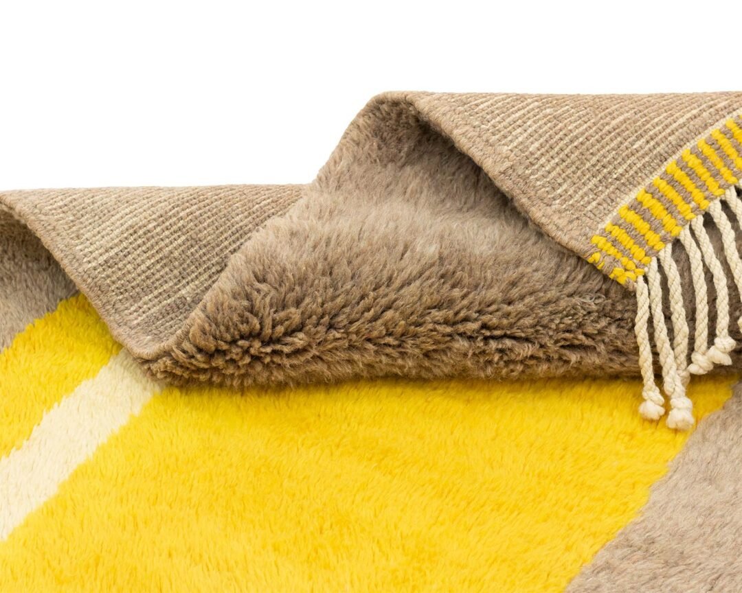This luxurious hand-knotted rug features a close-up of its folded design, showcasing a soft, fluffy texture in yellow and beige. The beige side reveals a flat weave accented with elegant yellow fringe.