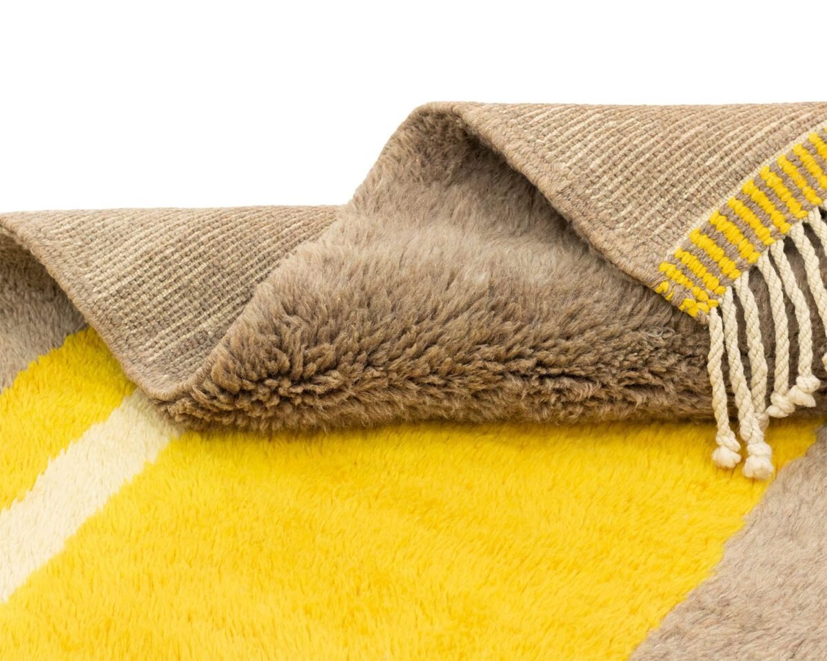 This luxurious hand-knotted rug features a close-up of its folded design, showcasing a soft, fluffy texture in yellow and beige. The beige side reveals a flat weave accented with elegant yellow fringe.
