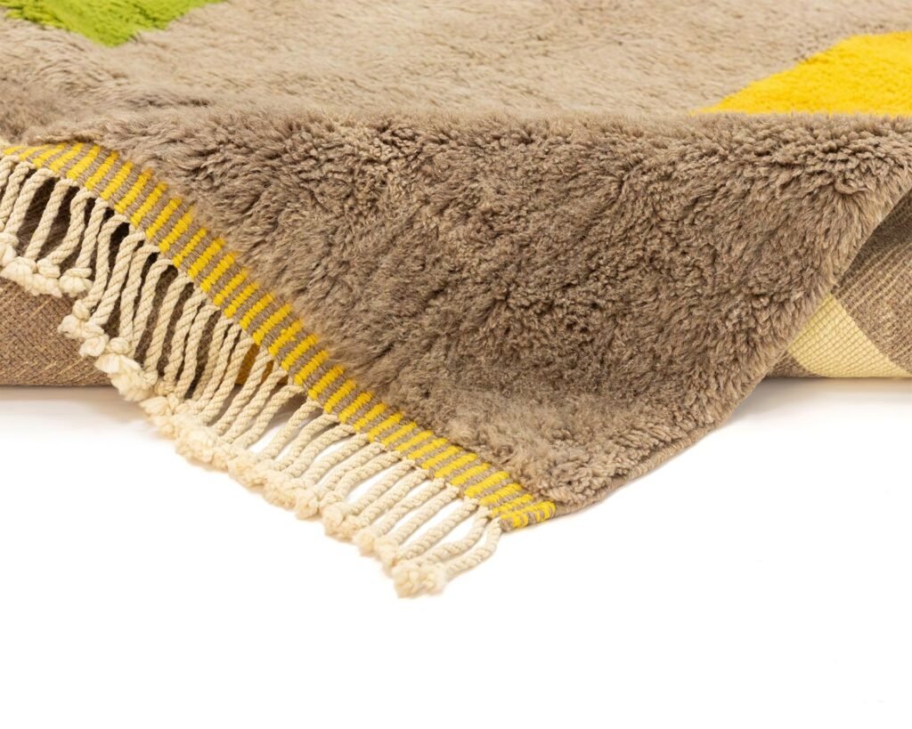Close-up of a fringed, hand-knotted Moroccan rug corner. The rug boasts a fluffy texture with rich brown, green, and yellow sections. White and yellow tassels elegantly hang from the edge, elevating its luxurious appeal.
