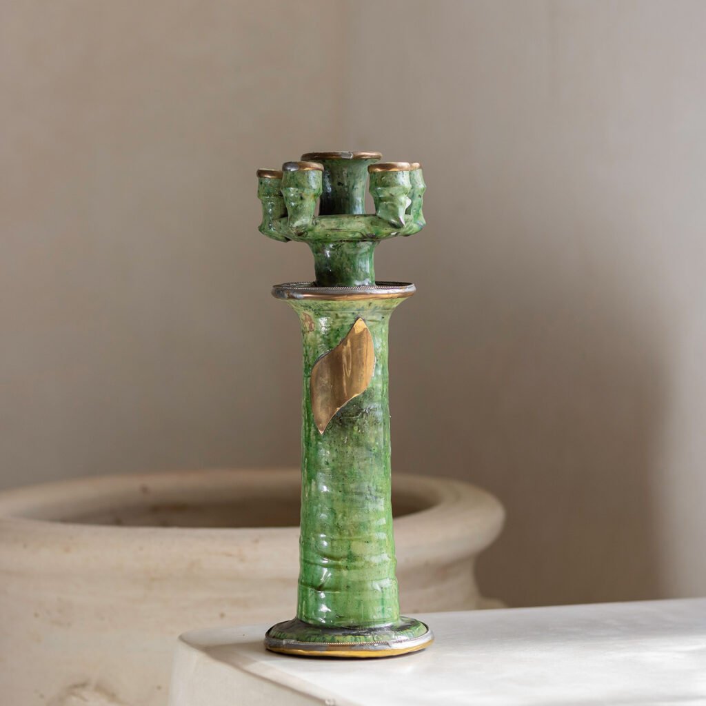 A green ceramic candle holder with gold accents, featuring multiple face sculptures around the top, sits elegantly on a light surface against a softly blurred background.