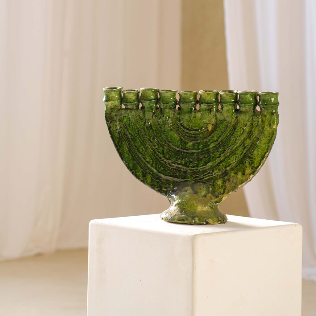 A stunning green ceramic menorah with nine candle holders elegantly displayed on a white pedestal, set against a warm beige background.