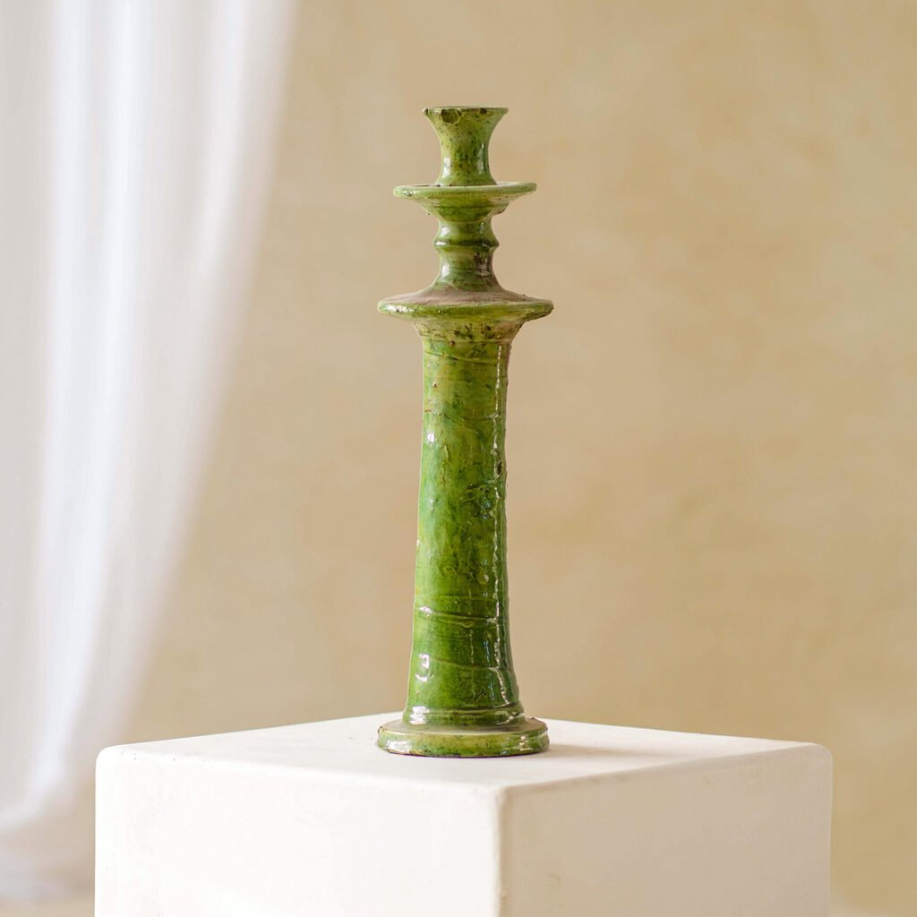 This elegant green ceramic candlestick boasts a glossy finish and a tall, slender design. Its beautifully showcased on a white square pedestal set against a soft, light backdrop.