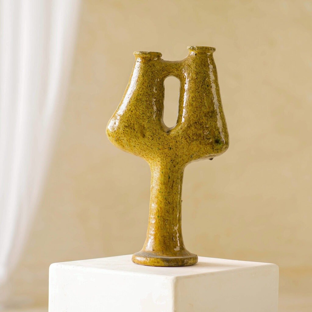 A textured, mustard-colored ceramic vase with three openings elegantly stands on a white pedestal against a neutral background, adding a captivating focal point to the space.